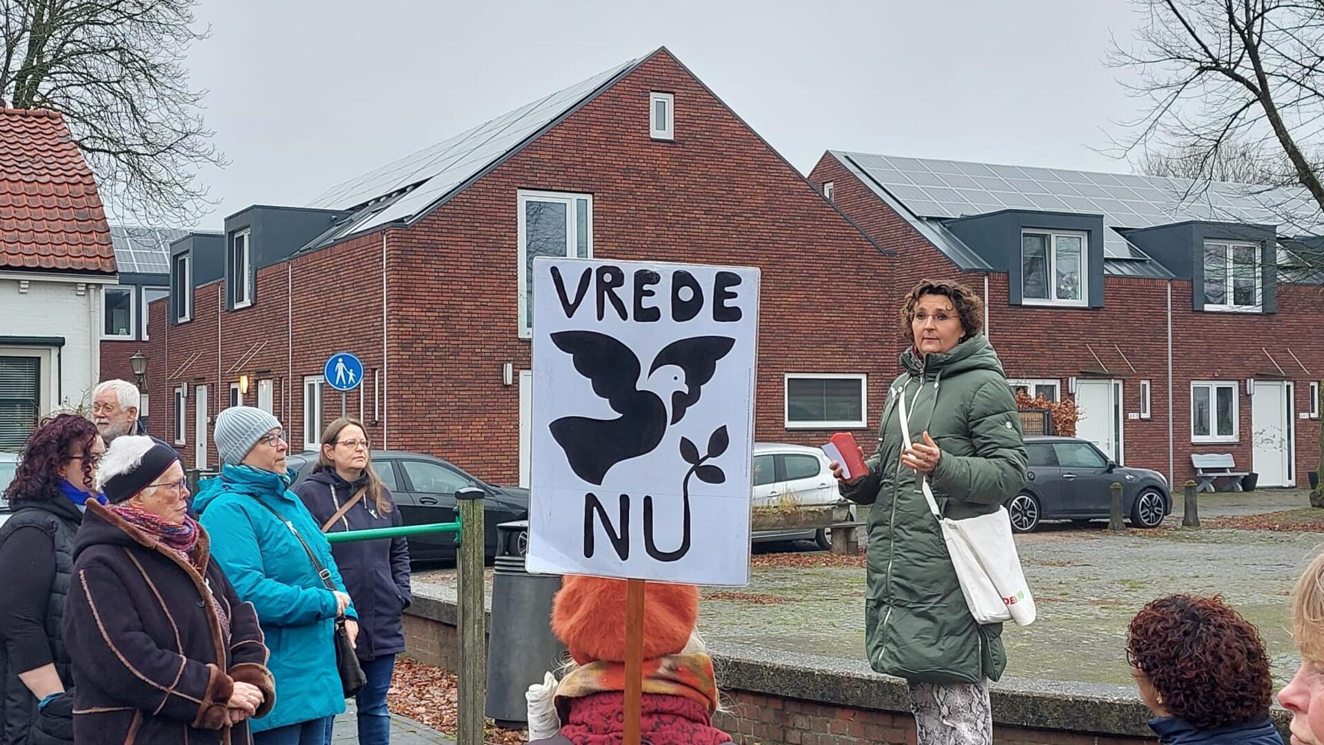 Een groep mensen staat op een plein. Een vrouw spreekt hen toe terwijl een deelnemer een bord met daarop de tekst: Vrede Nu omhoog houdt.